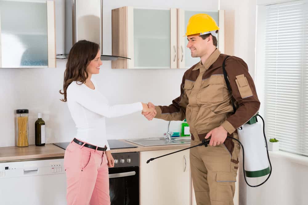 A woman in a white shirt and pink pants shakes hands with a man in a beige uniform and yellow hard hat inside a kitchen. The man holds a pest control spray tank on his back and a spray wand in his other hand. Kitchen cabinets and appliances are visible in the background.