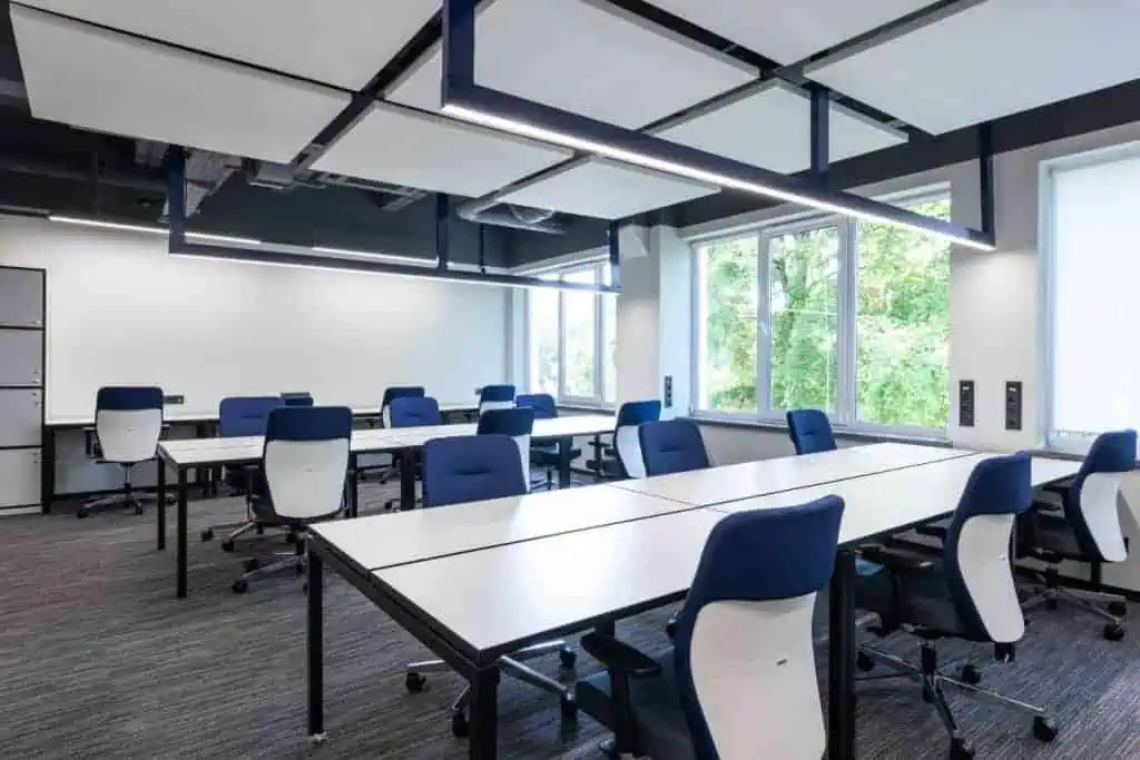 A modern office space with multiple workstations featuring blue and white chairs and white desks. Large windows provide natural light, and overhead lighting fixtures add brightness. Gray carpeting covers the floor.